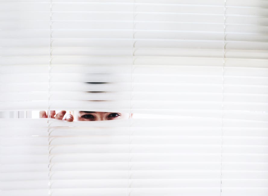 A person peeks through white blinds, creating an intriguing sense of curiosity.